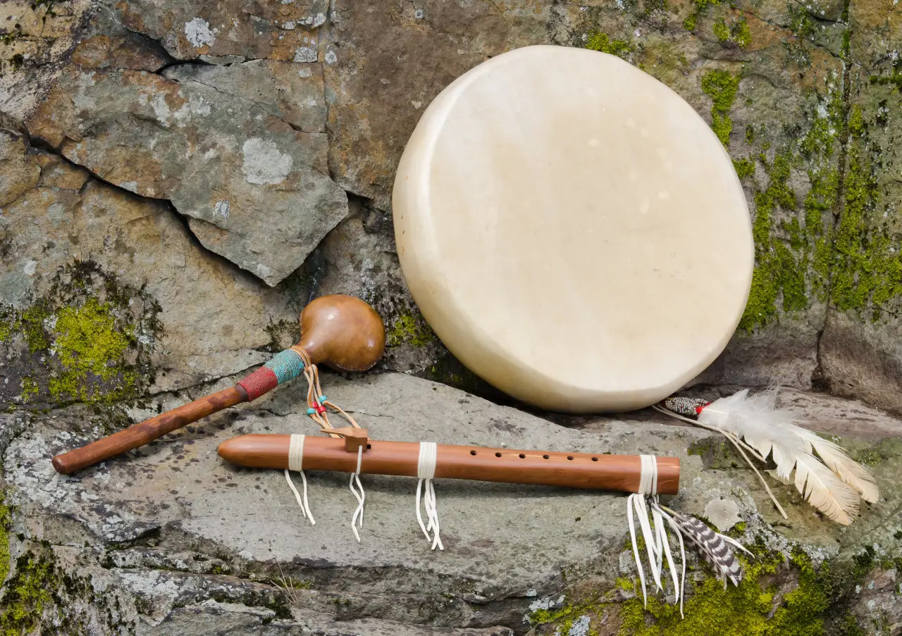 A picture of a large, flat, Native American Frame Drum with decorative feathers, and a Hand Carved Flute, and Shaker.